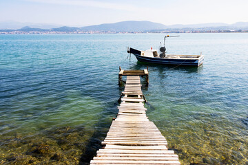Naklejka premium Traditional wooden pier and fishing boat on calm turquoise sea water with a coastal town and mountains in the background under a clear sky.