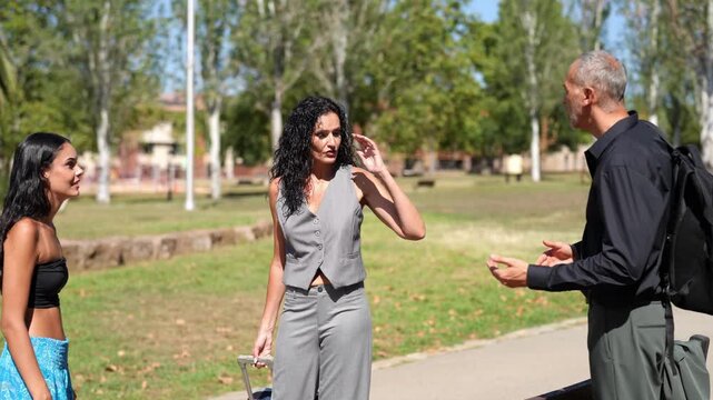 Sunlit park path with three people conversing. Woman in gray suit smiles, listens attentively. Other woman gestures with hand, engaged in talk. Man with backpack speaks, hands animated