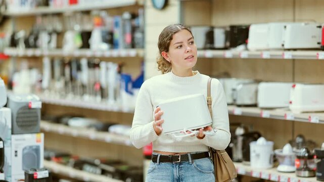 Young woman buyer examines toaster in detail before buying, checks completeness of household appliances and presence of defects. Customer chooses household appliances in store exhibition hall