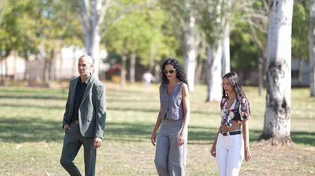 Three people stroll through a sunlit park. Trees stand tall, casting dappled shadows. One person wears a tailored suit, another a grey jumpsuit. Third wears a patterned top and white pants