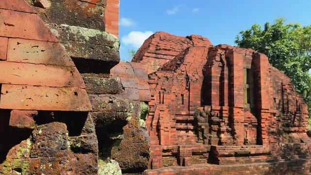 Cinematic pan shot revealing weathered red brick walls of ancient Cham temple ruins near Hoi An, Vietnam. Mossy stone textures and crumbling towers under blue sky.