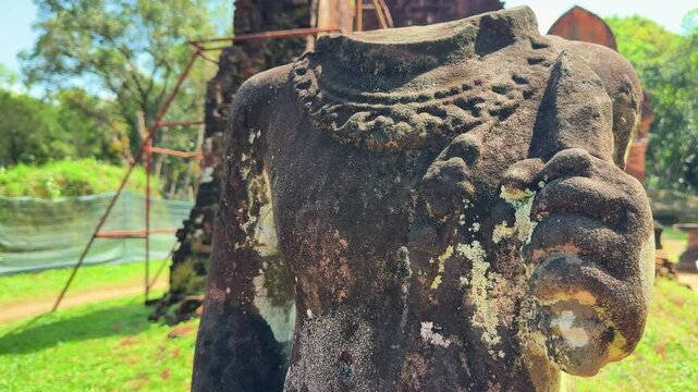 Weathered stone statue torso with intricate carvings at My Son Sanctuary, Hoi An, Vietnam. Ancient Cham ruins with moss-covered stonework and temple towers in background.