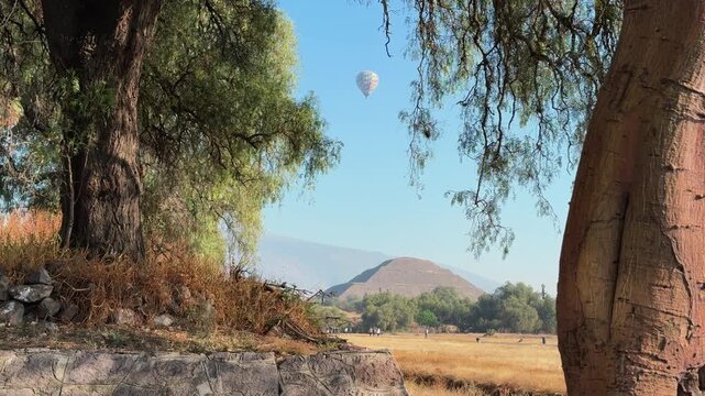Hot air balloon drifts peacefully over the ancient pyramids of Teotihuacan, Mexico, framed by lush trees under a clear blue sky.