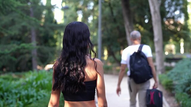 Young woman walks with long dark hair. Man walks ahead, carrying a backpack and suitcase. They stroll on a sunlit park path. Trees line the walkway, casting dappled shadows