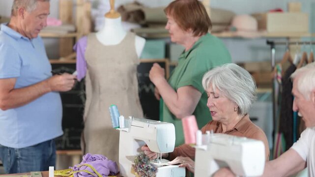Man and woman focusing on stitching at machines during group class for seniors in tailoring studio, while fellow learners preparing pattern pieces for assembly and adjusting fabric on mannequin