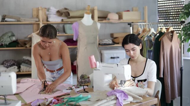  Team of dressmakers works in a sewing workshop. Fashion designer carefully cuts the fabric next to the assistant, who sews on the machine
