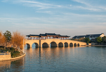 Obraz premium Covered stone arch bridge with multiple spans extends across a placid lake during sunset.
