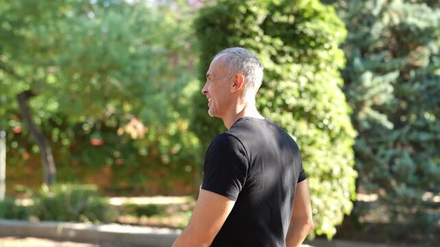 Man walks outdoors, smiling, relaxed. Green trees and bushes surround him. Sunlight filters through leaves behind him. He wears simple black t-shirt, appears content. Ideal for wellness, fitness