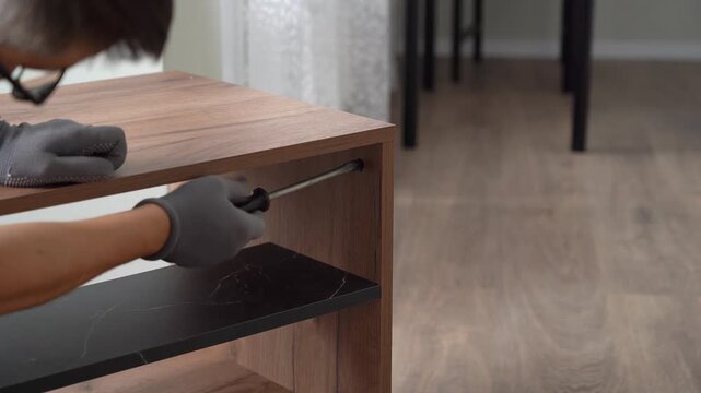 Furniture assembler assembles wooden nightstand using screwdriver. Process of assembling furniture