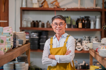 Fototapeta premium Proud senior Asian man in a yellow apron stands with arms crossed smiling in his beautiful ceramics and lifestyle shop.