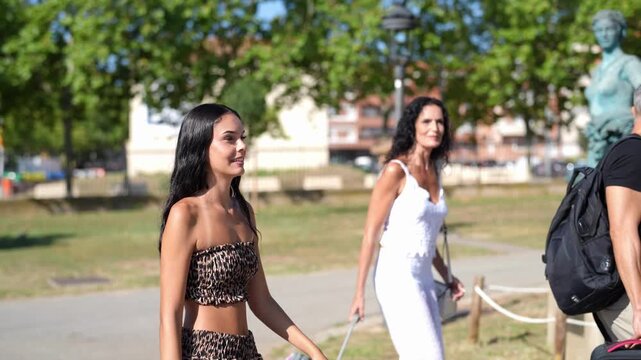 Two women stroll through a sunny park. One wears a leopard print two-piece outfit. Another wears a white dress, carrying a suitcase. They walk past statues and trees under bright daylight