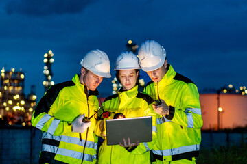 Fototapeta premium Petrochemical industry night engineer teamwork working on laptop Team of Petroleum in Glowing Workwear Collaborating oil refinery plant construction site at night
