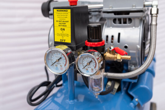 Close-up of an air compressor pressure switch assembly, featuring a black control box with a red emergency stop button, dual analog pressure gauges, and a red output pressure regulator knob