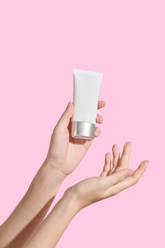 Two hands gently presenting cosmetic cream jar on pastel pink background. Clean minimal studio lighting with blank packaging. Cream product mockup and cosmetic promotion.