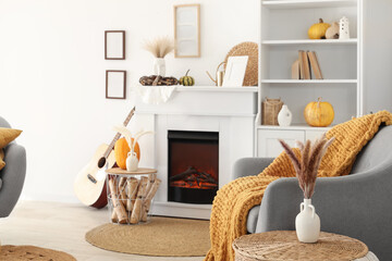 Obraz premium Interior of living room with armchair, coffee table, guitar, autumn home decor on fireplace and pumpkins in shelving unit near white wall