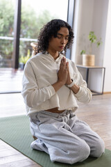 Fototapeta premium Nonbinary person kneeling on green yoga mat at home, wearing cream crop hoodie holding prayer hands