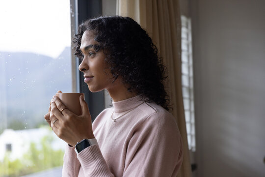 Non-binary adult standing near rain-speckled window holding ceramic mug, wearing pink sweater