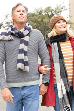 Middle-aged couple walking arm-in-arm on tree-lined urban sidewalk, wearing scarves and smiling