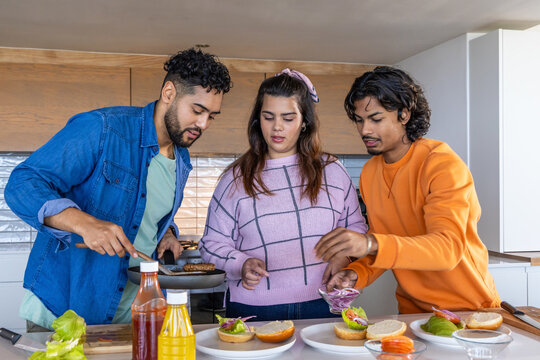 Indian friends cooking burgers at kitchen counter, flipping patties with spatula and topping buns