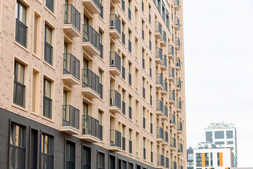 Fototapeta premium Modern apartment building stands tall against cloudy sky. Uniform balconies line beige brick facade with dark railings. Architectural pattern repeats vertically, creating rhythmic symmetry