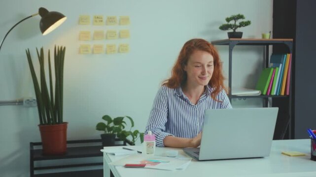 Energetic woman with fiery hair enjoys remote communication amid vibrant desk setup and lush greenery