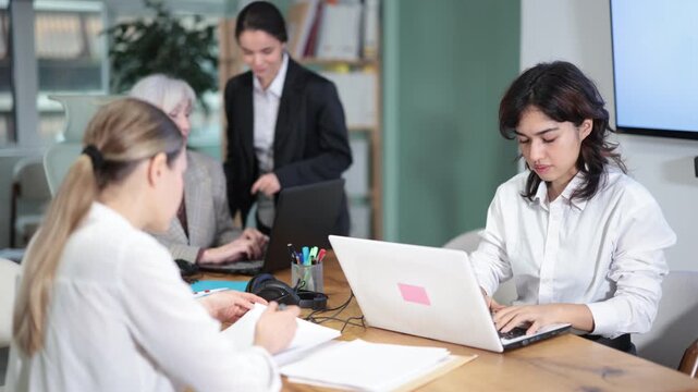 Asian girl sitting at desk is working on laptop computer among staff colleagues. Working day in office of business structure, manager is working at computer. 