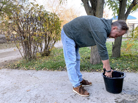 adult man bending over bucket and soaking sponge in water during manual car wash outdoors near private house autumn season everyday lifestyle vehicle care preparation step
