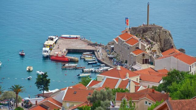 High angle view of Petrovac harbor in Montenegro, featuring traditional orange tiled roofs, a stone pier with boats, and the calm turquoise Adriatic Sea. A perfect summer travel and vacation scene
