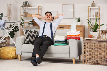 Obraz premium Handsome young happy businessman with iron and stacks of clothes sitting on sofa at home