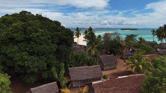 Top-down view of paradise beach. Palm fronds frame crystal clear water. Wooden boats float near sandy shore. Visitors sunbathe or paddle in calm ocean. Perfect for tourism, advertising