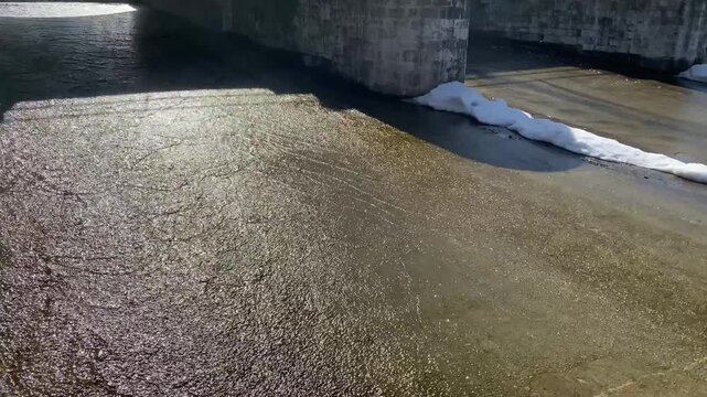 Cascading ponds connected by overflow weir. Water flows under bridge through stepped spillway in city park. Active melting of snow and ice. Spring flood.