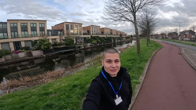 Young employee with shaved head vlogging while walking along a canal in a modern dutch neighborhood