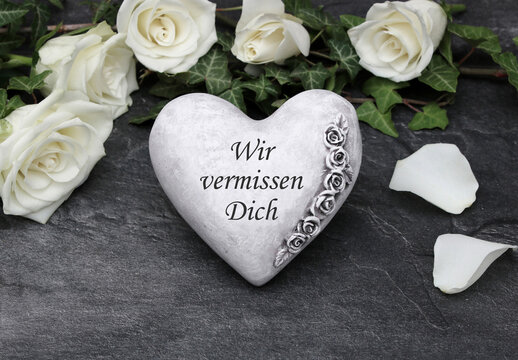 Trauerkarte oder Kondolenzkarte: W&uuml;rdevoller Abschied: Steinherz mit Schriftzug 'Wir vermissen Dich' umrahmt von wei&szlig;en Rosen und Efeu auf Schiefer.&ldquo;