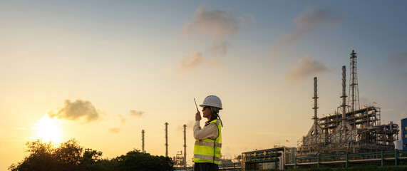Female petroleum engineer in safety helmet and vest using radio communication for talking project with tablet at oil refinery site during sunset engineering operation and energy industry. © BESTIMAGE