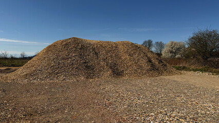 Naklejka premium Large mound of gravel and stones on a construction site under a clear blue sky with sparse vegetation and trees in the background