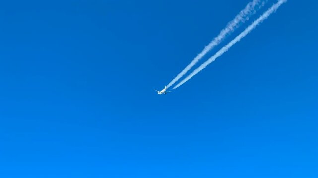 Aerial view of a twin-engine white color jet airplane and its fading contrail in a blue sky. Shot taken from a jet cockpit flying bellow.