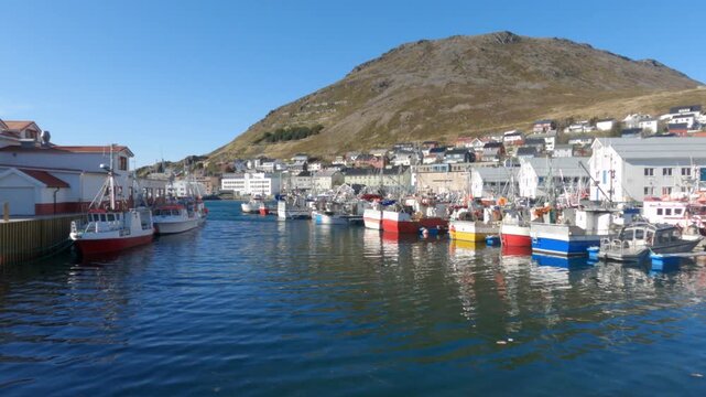 Scenic Norwegian Arctic harbor at Honningsv&aring;g near Nordkapp with colorful fishing boats moored along the quay, calm blue water, and rugged mountain backdrop under clear summer sky.