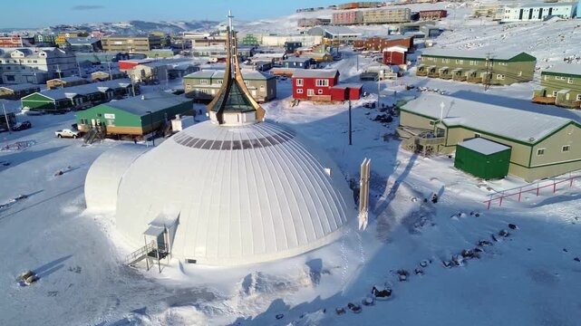 Drone flight over the iconic St. Jude's Cathedral, revealing the surrounding urban infrastructure, housing, and Qajuqturvik Food Centre in the background of the Arctic capital.
