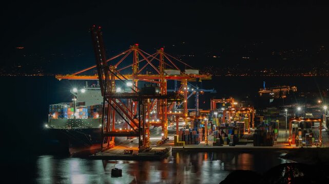Timelapse of quay and gantry cranes loading and unloading a suezmax container ship at a bustling international port. City lights in the background and reflections in the water.