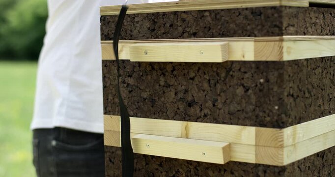 Close up of a hand tightening a black ratchet strap around a modern sustainable beehive made of cork and wood