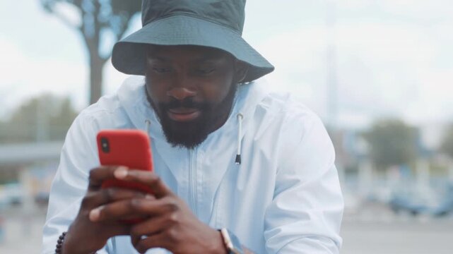 Casual man checking messages outdoors, Relaxed individual wearing bucket hat using phone near bus stop in city