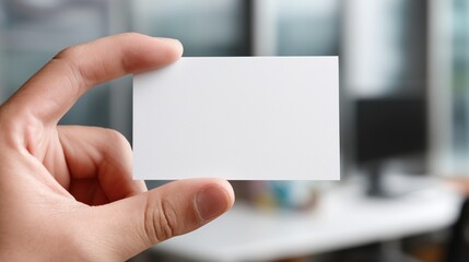 Obraz premium Hand holding a blank business card in an office setting with blurred background elements including a computer and desk