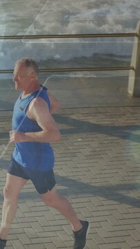 Vertical video: Senior man jogging by sea entering frame from left, passing railing for health