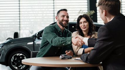 Couple discussing auto deal with salesman, shaking hands at a table inside a modern car dealership, vehicle visible in the background © wedmoments.stock