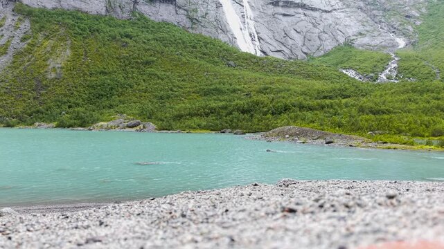 Aufnahme des Briksdalsbreen Gletschersees bei Regenschauer. t&uuml;rkises Wasser flie&szlig;t in einen Bach mit B&auml;umen im Hintergrund. Raw unbearbeitet