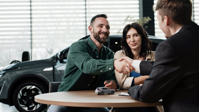 Couple discussing auto deal with salesman, shaking hands at a table inside a modern car dealership, vehicle visible in the background