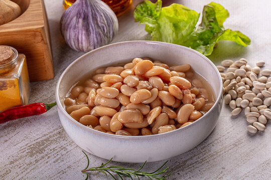 Bowl of cooked white beans surrounded by fresh ingredients like garlic, rosemary, and spices on a rustic table.