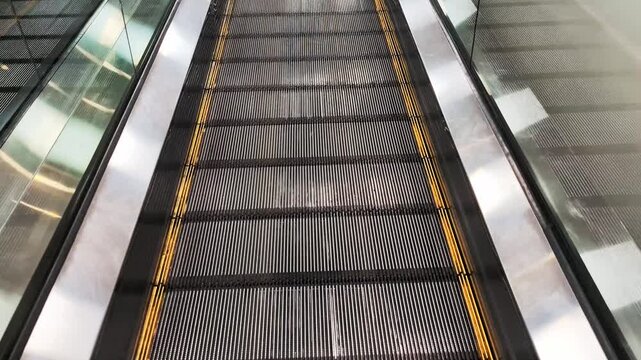 Point of view riding a moving escalator down inside a modern public building, with reflections on the glass and metal handrails providing an abstract urban perspective