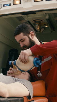Male emergency responder in red uniform administers oxygen to injured person using a bag valve mask inside an ambulance with medical equipment visible in the background