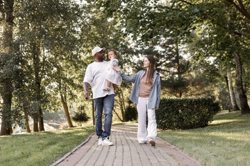 Fototapeta premium Happy interracial family walking in park on sunny summer day. Father holding little daughter in arms while mother walks. Warm lifestyle, parenting outdoors. People walking